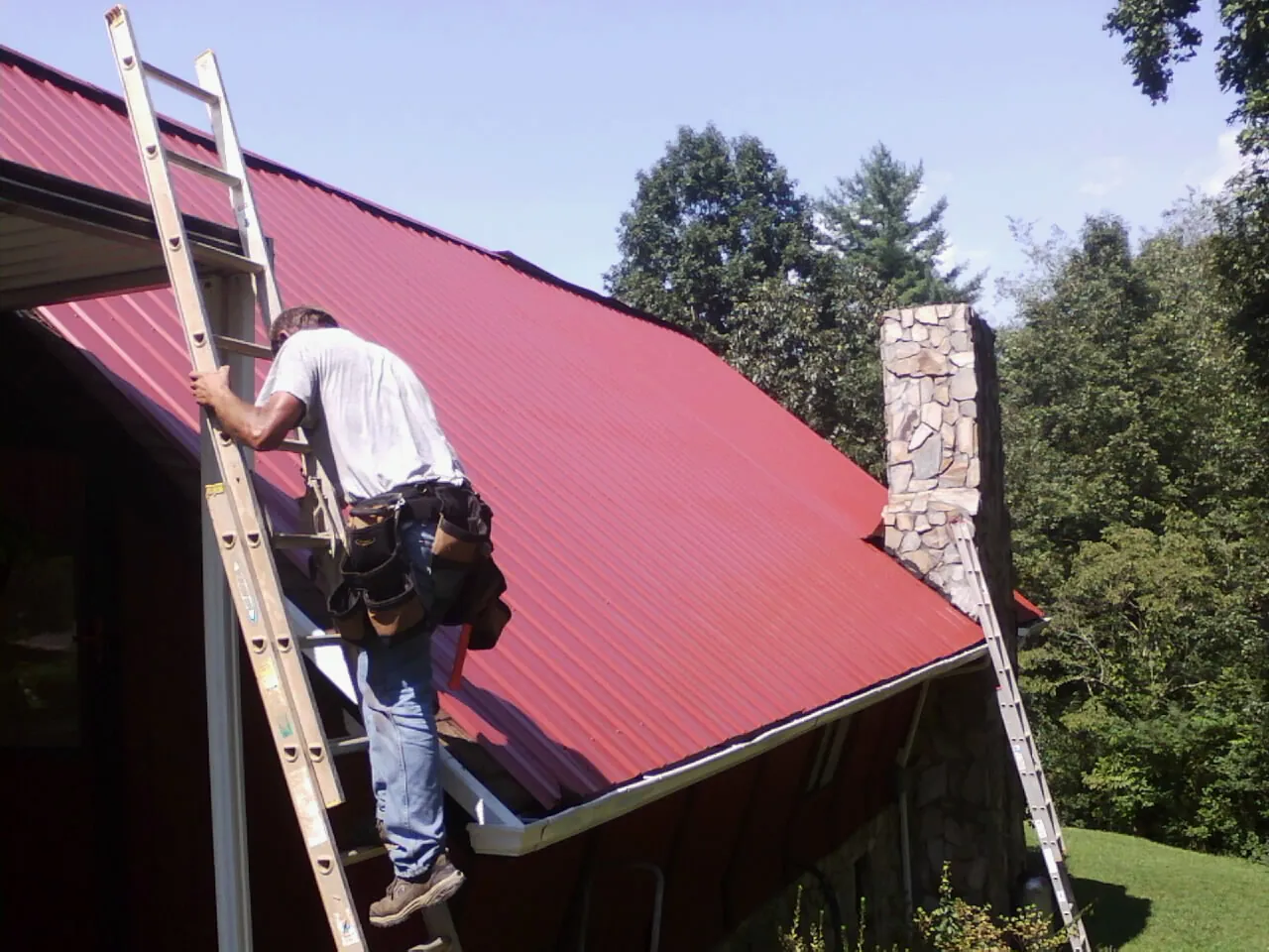 Roofer on a ladder beside a red metal roof and stone chimney — Soffit & Fascia Repair by Roofing Brothers in Broxton